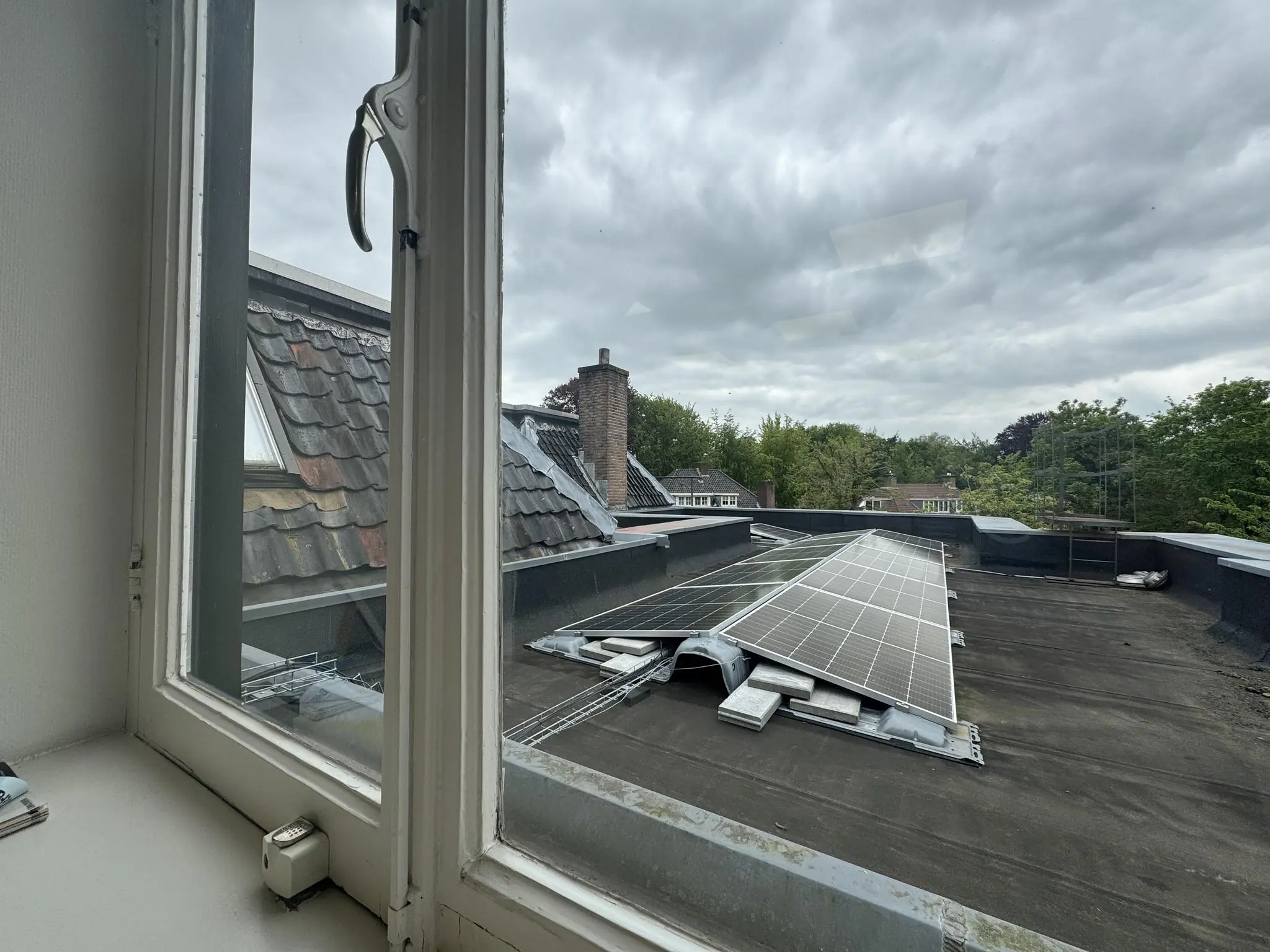 Uitzicht vanuit een raam op een plat dak met zonnepanelen aan de Dorpsstraat ter hoogte van de Steenstraat, omringd door bomen en aangrenzende daken.